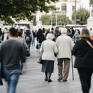 Coppia di anziani che cammina in una strada cittadina affollata, immagine che richiama l’invecchiamento della popolazione e i cambiamenti demografici.